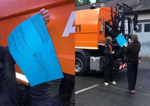 Zwei Personen präsentieren ein blaues Plakat vor einem orangefarbenen städtischen Reinigungsfahrzeug, während eine von ihnen es fotografiert.
