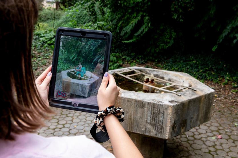 Eine Person hält ein Tablet vor einen alten Brunnen, auf dem Bildschirm wird durch Augmented Reality eine Szene mit zwei virtuellen Figuren über dem realen Brunnen dargestellt.