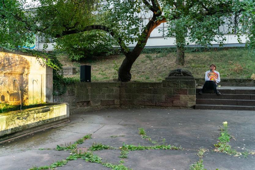 Das Bild zeigt eine Person, die auf einer Treppe in einem verwilderten Parkbereich sitzt und in ein Mikrofon spricht. Neben ihr steht ein Glas Weißwein. Im Hintergrund ist ein Brunnen mit gelblicher Beleuchtung zu sehen.