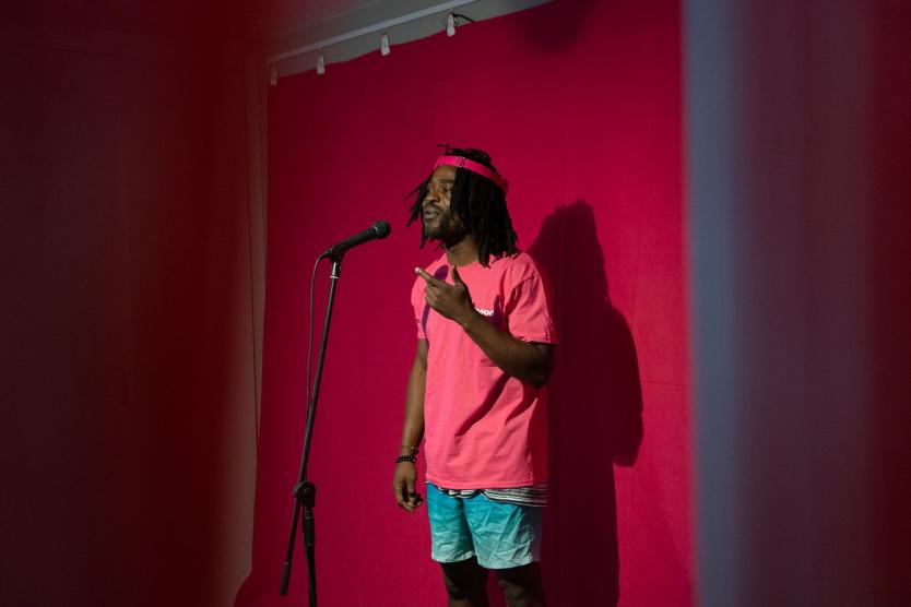 Das Bild zeigt einen Mann, der vor einem Mikrofon steht und mit einer Handgeste hinein spricht; hinter ihm hängt ein pinker Vorhang an der Wand.