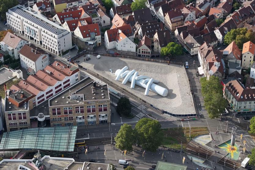 Das Bild zeigt eine Luftaufnahme eines städtischen Platzes, auf dem eine große weiße, aufblasbare Skulptur in einem leeren Gelände platziert ist. Umgeben ist der Platz von roten Ziegeldachhäusern, Geschäften und Straßenverkehr.