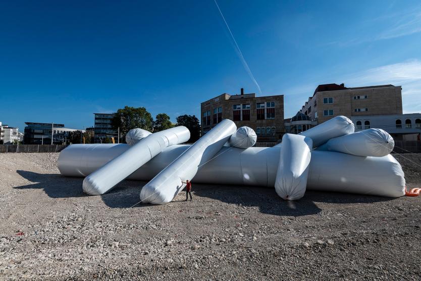 Das Bild zeigt eine eingezäunte Baugrube, in deren Zentrum eine monumentale Installation in Form eines Mikroorganismus aus aufgeblasenen Luftschlangen liegt. Ein Mann, der im Verhältnis zur imposanten Struktur klein erscheint, steht davor und berührt eine der Schlangen.