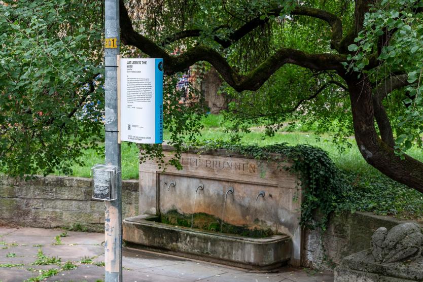 Das Bild zeigt einen historischen Brunnen mit drei Wasserspeiern, umgeben von dichten grünen Bäumen. Vor dem Brunnen hängt ein Schild mit der Aufschrift „JUST LISTEN TO THE WATER“ und weiteren Informationen, die zur Kunstinstallation gehören.