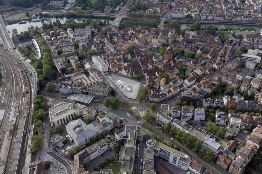 Das Bild zeigt eine weitläufige Luftaufnahme einer Stadt mit einer aufblasbaren weißen Skulptur im Zentrum eines großen Platzes. Rundherum sind Wohnhäuser, Straßen, Bahngleise und eine Brücke über einen Fluss zu sehen.