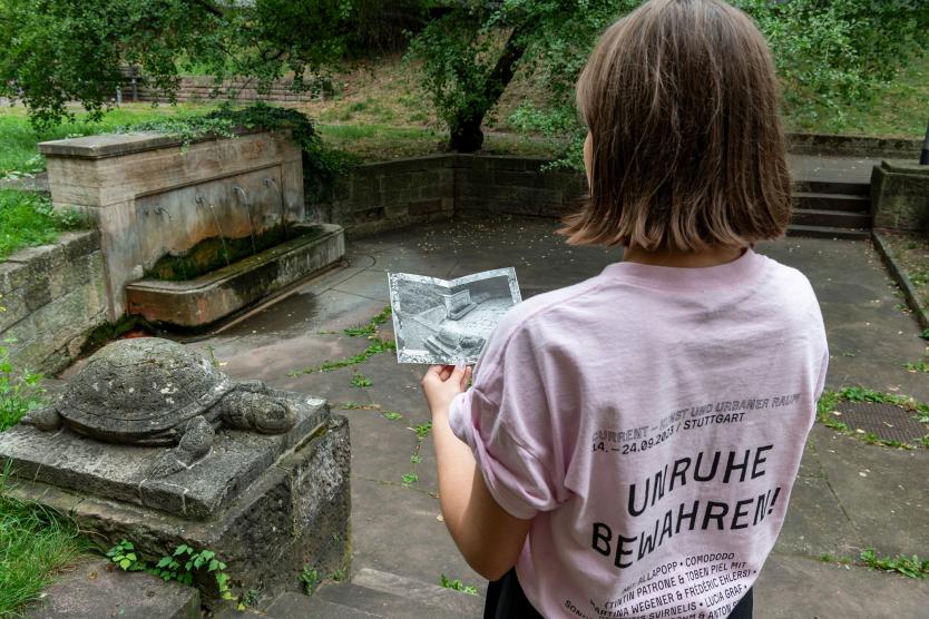 Eine Person mit rosa T-Shirt, auf dessen Rücken „UNRUHE BEWAHREN!“ steht, betrachtet ein Schwarz-Weiß-Foto, das den Brunnen mit einer Steinschildkröte zeigt. Der Brunnen ist mit Moos überwuchert, während die grüne Umgebung und Steinwände den Ort ruhig wirken lassen.