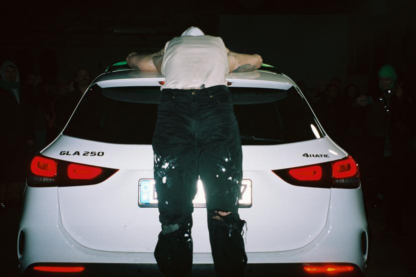 Das Bild zeigt eine Person mit weißem T-Shirt und verschleiertem Kopf, die bäuchlings mit ausgestreckten Armen auf dem Dach eines weißen Autos liegt, während im Hintergrund nur das Fahrzeug und die dunkle Umgebung zu sehen sind.