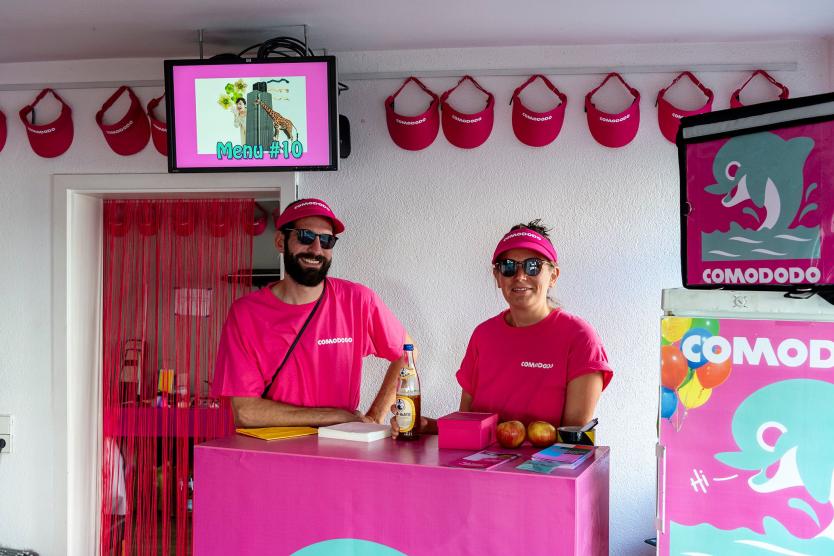 Das Bild zeigt zwei Personen, die hinter einer Theke stehen. Rechts von ihnen sind Werbeartikel zu sehen, oberhalb ein Bildschirm, der ebenfalls für den Lieferdienst wirbt. Links führt ein Gang in eine andere Räumlichkeit. Die Personen tragen Capes und Sonnenbrillen und lachen in die Kamera.