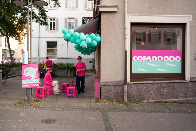 Das Bild zeigt den Vorplatz des Ladens mit fünf pinken Hockern, auf denen eine Person sitzt und eine Person steht. Über dem Eingang hängen grüne Ballons, und an dem Ladenfenster sowie an einer Straßensäule hängt ein Plakat, das für den Lieferdienst wirbt und einen Delfin zeigt.