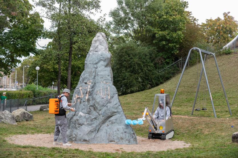 Das Bild zeigt zwei Personen vor einem künstlichen, grauen Felsen in einem Park. Einer putzt mit einem Tuch den Felsen, während der andere in einer kleinen, futuristischen Reinigungsmaschine mit blauen Bürsten sitzt. Die Szene spielt in einer grünen Umgebung mit Bäumen und Wiesen.