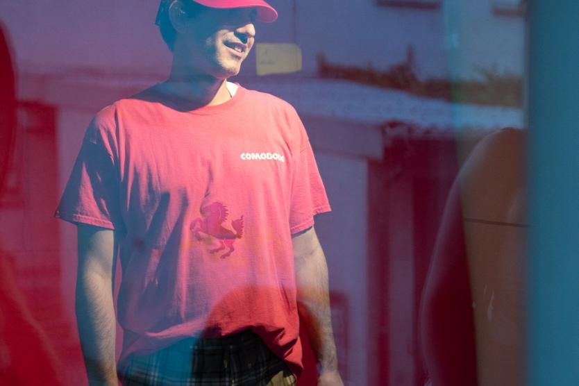 Das Bild zeigt einen Mann mit einem Mikrofon in einem Schaufenster stehen. Der Mann trägt ein Pinkes T-Shirt und eine Capie auf dem steht Comododo