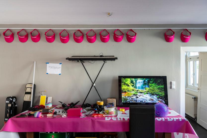 Das Bild zeigt eine lange Tafel, die an einer Wand in einem Raum steht und über der eine pinke Tischdecke liegt. Auf der Tafel stehen ein Klavier, verschiedene Süßigkeiten und ein Bildschirm. An der Wand oben hängen pinke Caps des Lieferdienst-Ladens.