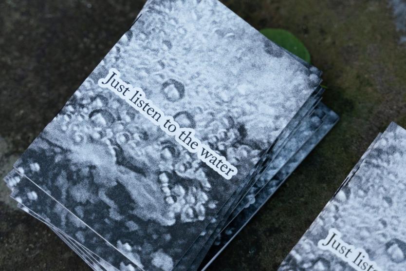 Das Bild zeigt einen Stapel schwarz-weißer Flyer mit der Aufschrift „Just listen to the water“. Die Struktur des Wassers ist als Hintergrundmotiv auf den Flyern gedruckt.