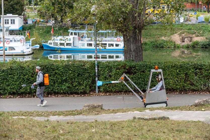 Das Bild zeigt eine Person, die auf einem Gehweg entlang eines Flusses läuft, während ein motorisiertes Reinigungsfahrzeug mit blauer Bürste hinter ihr fährt. Im Hintergrund spiegeln sich Boote im Wasser.