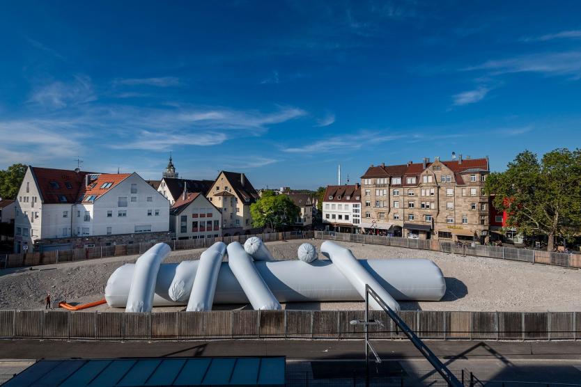 Das Bild zeigt das aufblasbare Kunstwerk aus der Ferne, umgeben von historischen Gebäuden und Bauzäunen auf einem offenen, kiesbedeckten Platz.