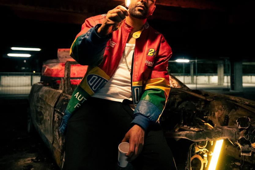 Das Bild zeigt den Künstler Bobby Sayyar, der in einem urbanen Setting sitzt. Die Lichtverhältnisse und seine Kleidung verleihen der Szene eine atmosphärische, kontrastreiche Stimmung.