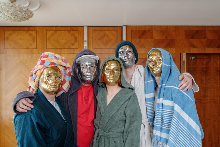 Das Bild zeigt fünf Personen, die in Bademänteln und mit Handtüchern auf dem Kopf posieren, während sie goldene und silberne Masken tragen; sie stehen eng zusammen vor einer hölzernen Wand mit geometrischem Muster.