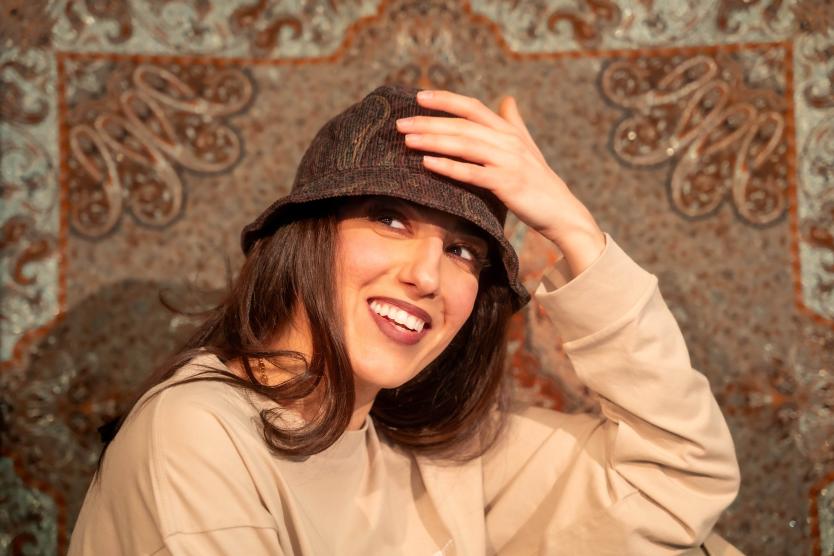 Das Bild zeigt die Künstlerin Donya Ahmadifar, in einem lässigen und warmen Ambiente. Sie lacht und hält sich leicht an ihrem Hut. Mit einem freundlichen Lächeln schaut sie in die Kamera.
