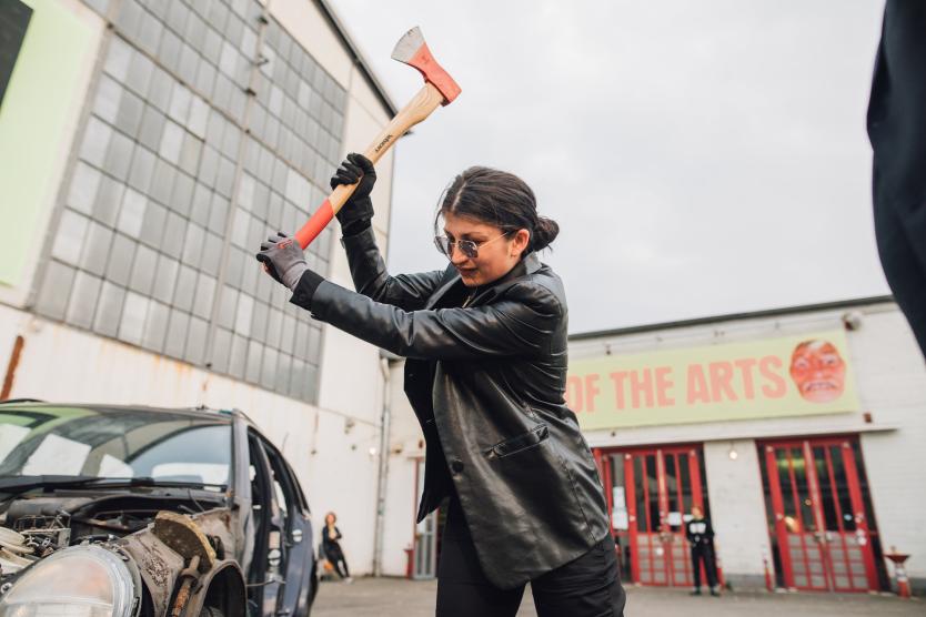 Das Bild zeigt dieselbe Person mit schwarzer Lederjacke, die eine Axt mit roten Details über einem Auto erhebt, bereit zuzuschlagen, vor einer Industriehalle mit der Aufschrift „STATE OF THE ARTS“.