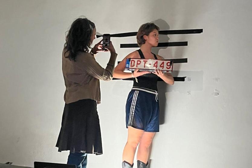 Das Bild zeigt eine Person, die mit kurzen Hosen und Cowboystiefeln vor einer weißen Wand steht, ein Nummernschild mit der Aufschrift „DPT-449“ hält und von einer anderen Person mit einem Smartphone fotografiert wird.