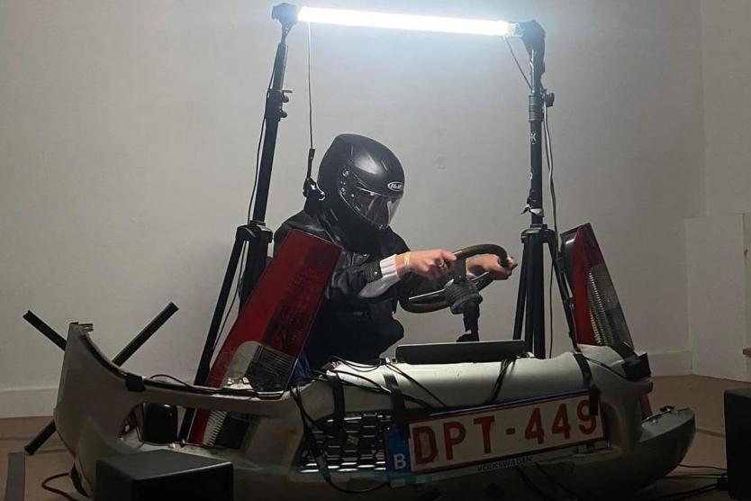 Das Bild zeigt eine Person mit Motorradhelm und Lederjacke, die auf einem improvisierten Fahrsimulator aus Autoteilen mit dem Nummernschild „DPT-449“ sitzt, umgeben von Licht und technischen Geräten.