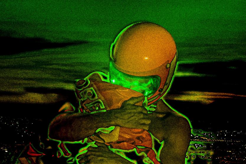 Das Bild zeigt eine Person mit orangefarbenem Helm und leuchtend grünem Visier, die ein oranges Paket fest umarmt, vor einem grünlich verfremdeten nächtlichen Stadtpanorama.