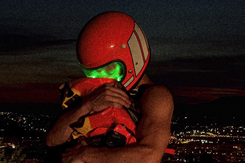 Das Bild zeigt eine Person mit orangefarbenem Helm und leuchtend grünem Visier, die ein oranges Paket fest umarmt, vor einer nächtlichen Stadtlandschaft mit leuchtenden Lichtern im Hintergrund.