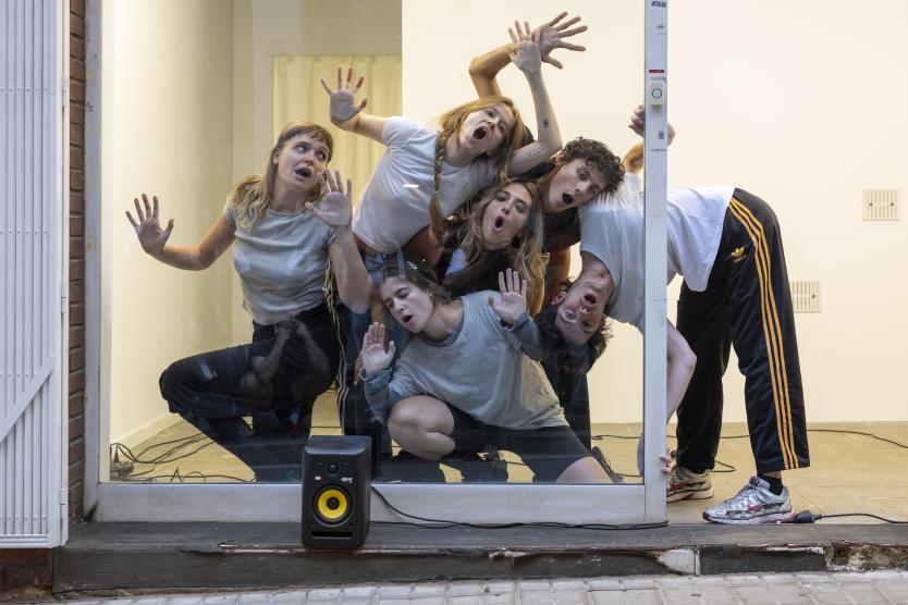 Das Bild zeigt mehrere Menschen, die sich von innen gegen die Glasscheibe eines Ladens drücken, wobei ihre Gesichter und Gliedmaßen am Glas haften.