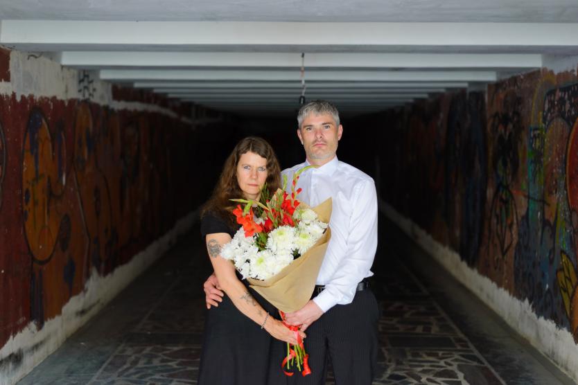 Das Bild zeigt ein Paar oder zwei zusammenstehende Personen in eleganter Kleidung, die sich mit ernsten Gesichtern in einem graffiti-bedeckten, dunklen Tunnel umarmen und einen großen Blumenstrauß mit weißen und roten Blumen halten.