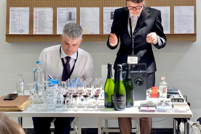 Das Bild zeigt zwei Personen in formeller Kleidung, die an einem Tisch voller Gläser, Champagnerflaschen und technischen Geräten sitzen, während sie konzentriert arbeiten; eine Person steht gestikulierend, während die andere etwas notiert.