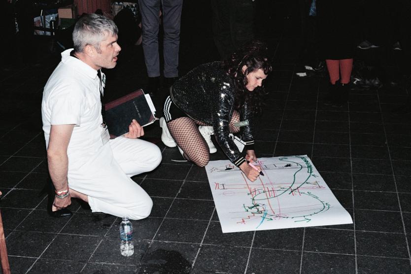 Das Bild zeigt eine Person in weißem Overall, die kniend mit einer anderen Person in glitzerndem Outfit und Netzstrümpfen interagiert, während diese eine große Zeichnung mit farbigen Linien und Symbolen auf dem Boden anfertigt.