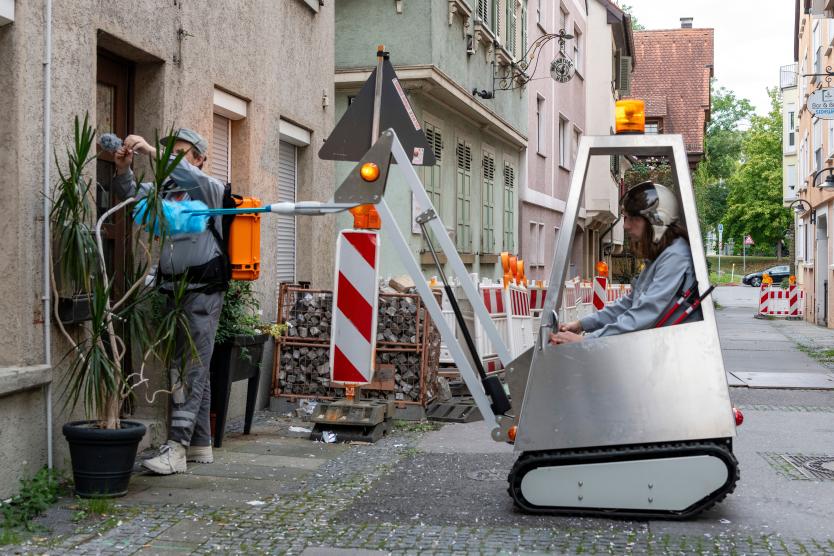 Das Bild zeigt eine Person in einem futuristischen Mini-Fahrzeug und eine andere Person, die mit einem verlängerten Reinigungswerkzeug eine Hausfassade säubert.