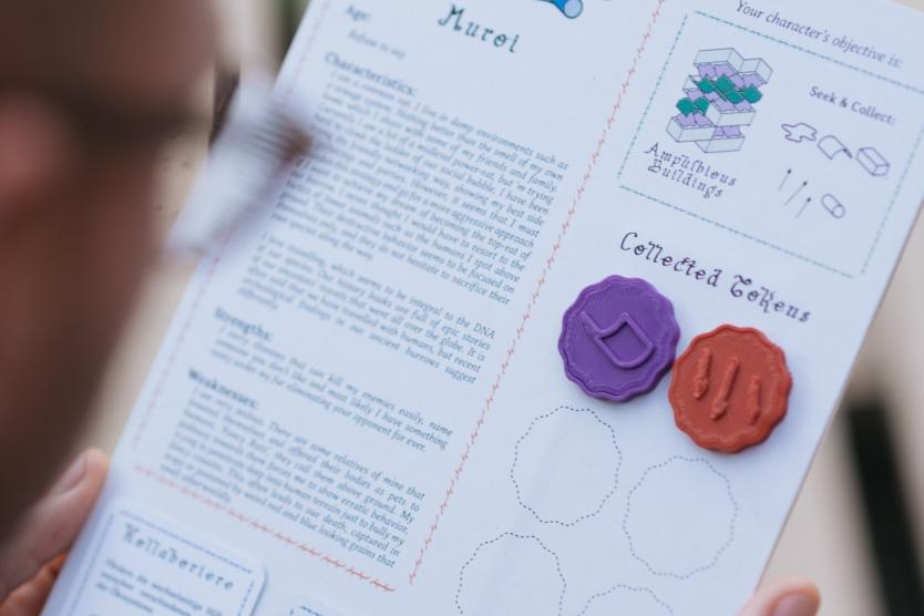 Das Bild zeigt ein handgefertigtes Spielbrett mit Texten, Illustrationen und farbigen Token, die gesammelt werden; eine Person hält es mit beiden Händen und liest den Text.