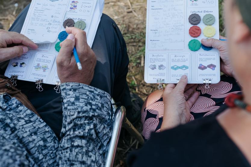 Das Bild zeigt zwei Personen, die jeweils ein handgefertigtes Spielbrett auf ihrem Schoß halten, während sie draußen auf Stühlen sitzen und Notizen machen.