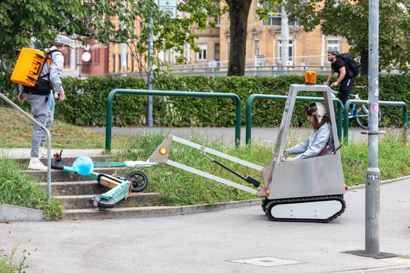 Das Bild zeigt eine Person, die eine Treppe hinaufgeht, während ein motorisiertes Reinigungsfahrzeug einen E-Scooter mit seiner blauen Bürste putzt. Ein Fahrradfahrer fährt im Hintergrund vorbei.