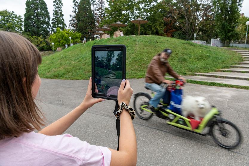 Eine Person sieht auf einem Tablet eine digitale Figur auf einer Treppe, während real ein Mann mit Hund auf einem Lastenrad vorbeifährt.