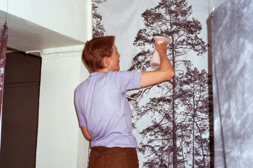 Eine Person steht mit dem Rücken zur Kamera und glättet mit einem Dampfbügeleisen ein großes Wandbild, das hohe Bäume zeigt. Die Szene spielt in einem Innenraum, vermutlich in einer Ausstellung.