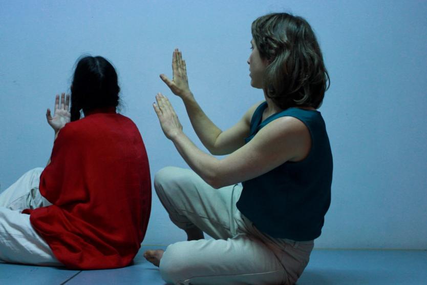 Zwei Personen sitzen barfuß auf dem Boden, mit dem Rücken zur Kamera. Beide haben die Hände vor sich erhoben, als würden sie eine imaginäre Wand ertasten. Die Lichtstimmung ist kühl, die Wand im Hintergrund hellblau.