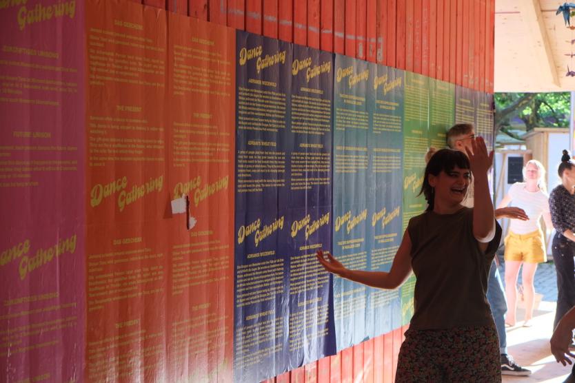 Tanzende Menschen vor einer bunten Wand mit „Dance Gathering“-Postern in einem sonnigen, überdachten Außenbereich.