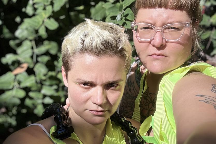 Zwei Personen blicken ernst in die Kamera. Sie tragen helle Oberteile und neongelbe Gurte um den Hals. Im Hintergrund sind große, grüne Blätter im Sonnenlicht zu sehen.