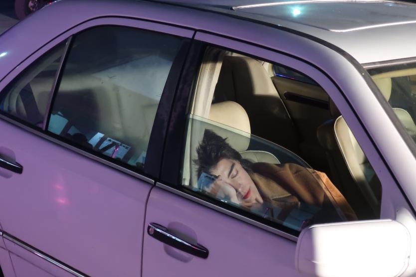 Das Bild zeigt eine Person mit geschlossenen Augen, die auf dem Rücksitz eines geparkten Autos liegt und sich mit dem Kopf an die Fensterscheibe lehnt