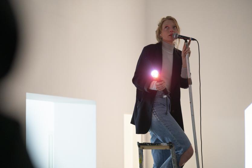 Eine Person steht auf einer Leiter, hält ein Mikrofon in der einen Hand und eine leuchtende Lampe in der anderen. Sie trägt Jeans, Rollkragenpullover und einen dunklen Blazer. Der Raum ist hell und minimalistisch.