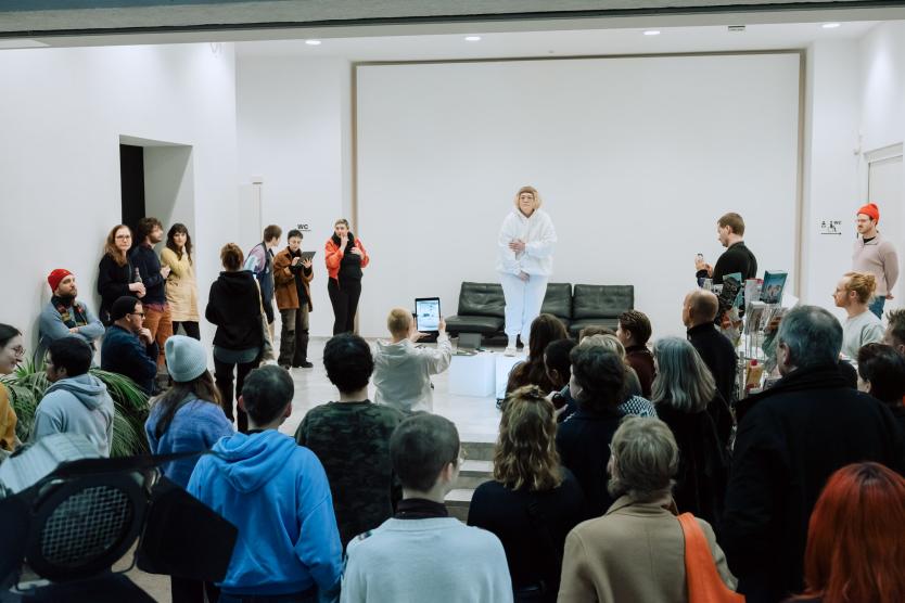 In einem weißen Ausstellungsraum steht eine Person in weißer Kleidung auf einer kleinen Plattform und performt. Rundherum schaut ein Publikum zu, einige filmen mit Mobilgeräten.