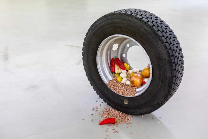 Das Bild zeigt einen aufrecht stehenden Autoreifen, dessen Felge mit Lebensmitteln wie Zwiebeln, Knoblauch, Paprika und Getreide gefüllt ist, wobei einige der Körner auf den Boden rieseln.