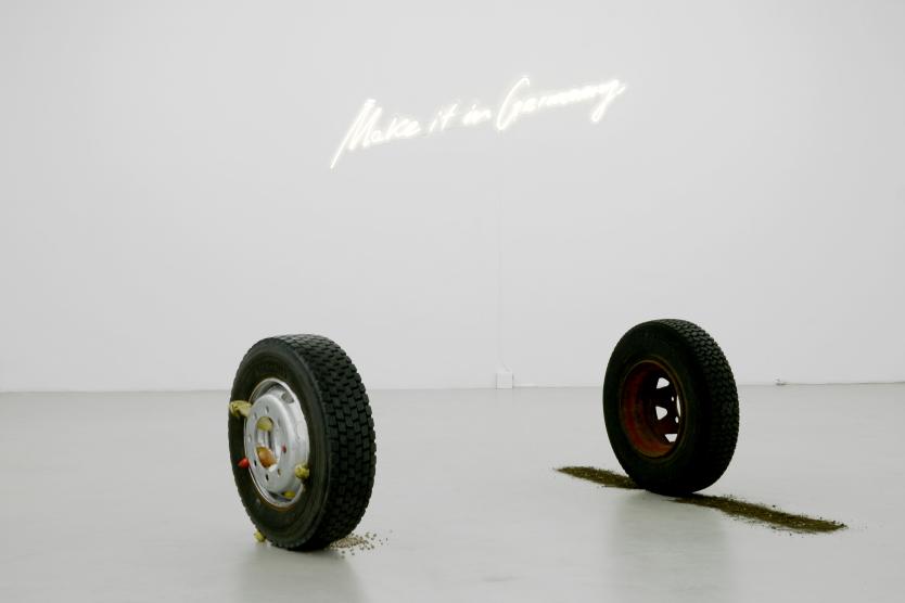 Das Bild zeigt eine minimalistische Kunstausstellung mit zwei stehenden Autoreifen, aus denen Erde und Lebensmittel herausquellen, vor einer weißen Wand mit der leuchtenden Schrift „Make it in Germany“.