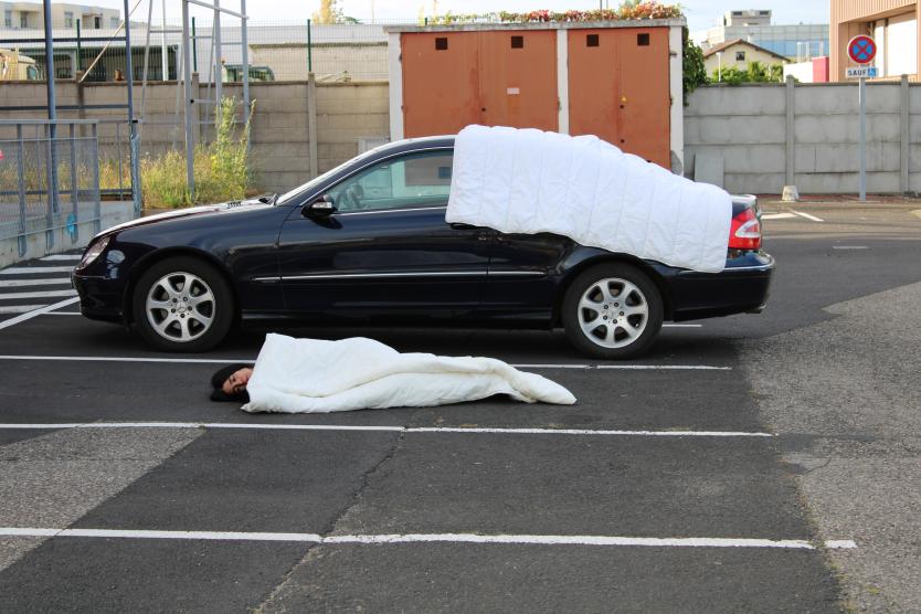 Das Bild zeigt eine Person, die in eine weiße Decke gewickelt auf einem Parkplatz vor einem schwarzen Auto liegt, auf dem ebenfalls eine weiße Decke ausgebreitet ist.