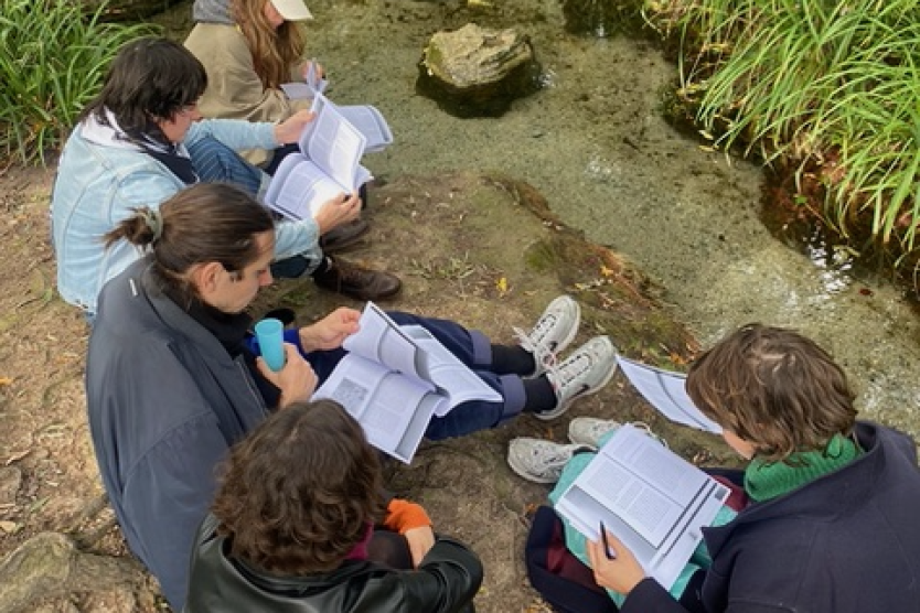 Das Bild zeigt eine Gruppe von fünf Personen, die am Ufer eines kleinen Baches im Wald sitzen, vertieft in Bücher und Notizen, während sie von dichter Vegetation umgeben sind.