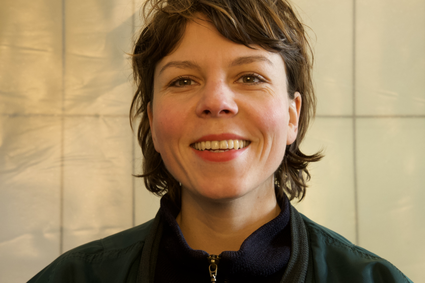 Eine lächelnde Frau steht vor einer hellen Wand und schaut direkt in die Kamera.