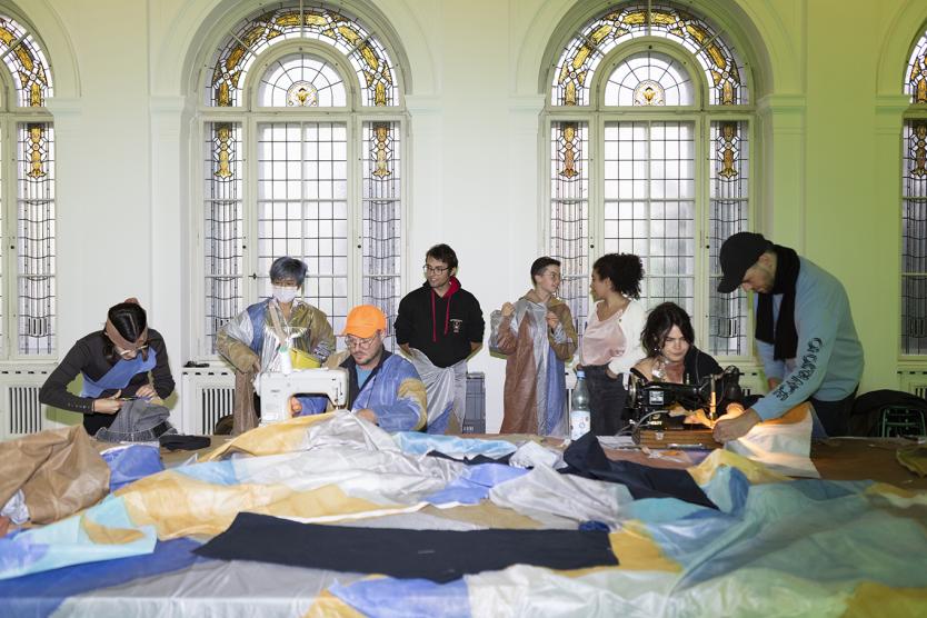 Das Bild zeigt mehrere Personen, die in einem hellen Raum gemeinsam an einer großen textilen Fläche nähen, schneiden und arbeiten.