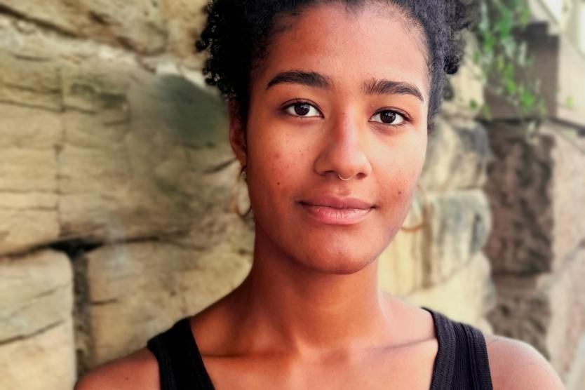 Porträt einer jungen Frau mit dunkler, lockiger Hochsteckfrisur. Sie trägt ein schwarzes ärmelloses Oberteil und blickt ruhig in die Kamera. Im Hintergrund ist eine Natursteinmauer mit etwas grünem Efeu zu sehen, die Szene ist bei Tageslicht aufgenommen.