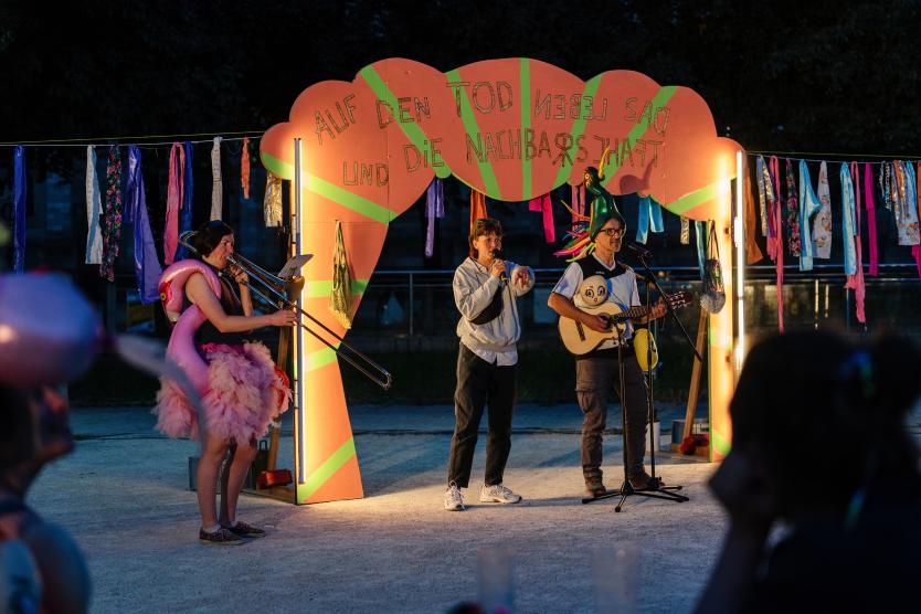 Drei Künstler:innen führen bei einem bunten Open-Air-Theaterstück unter einem leuchtenden Torbogen auf.